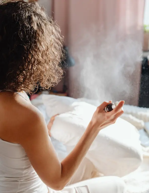 Person with curly hair holding a spray bottle, releasing a fine mist in a cozy bedroom setting with sunlight streaming in, suggesting a relaxing or self-care moment.