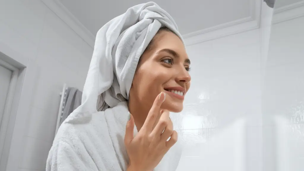 Smiling woman in a bathroom, wearing a white towel wrapped around her hair and a white robe, applying skincare product to her cheek with her fingertip.