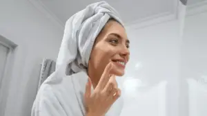 Smiling woman in a bathroom, wearing a white towel wrapped around her hair and a white robe, applying skincare product to her cheek with her fingertip.