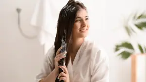 Smiling woman in a white robe applying a hair treatment mask to her long, dark hair in a bright bathroom with white walls and green plants in the background.