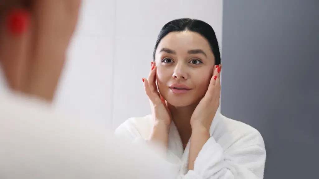 Woman with dark hair and red nails gently touching her face while looking into a mirror, wearing a white robe in a bathroom setting