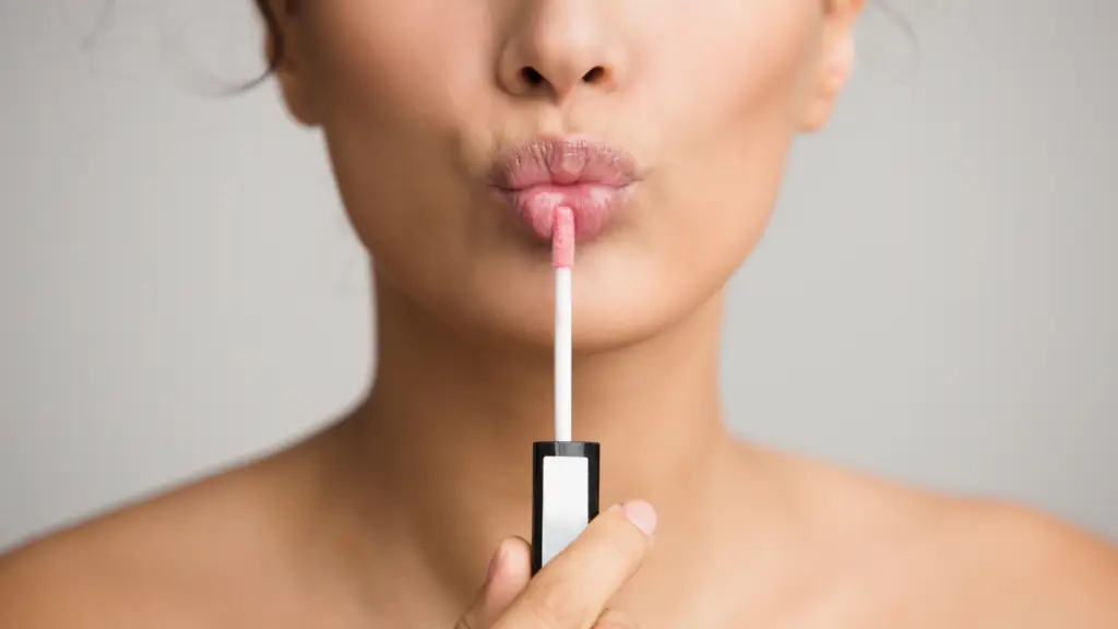 Close-up of a person applying pink lip gloss with an applicator, lips slightly puckered, focusing on the smooth, glossy finish.