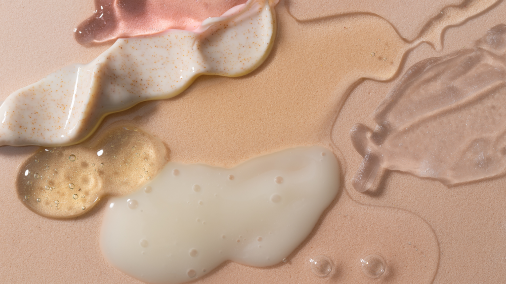 Close-up of various skincare product textures, including creams, gels, and oils, in shades of white, pink, and translucent with visible bubbles and exfoliating beads.