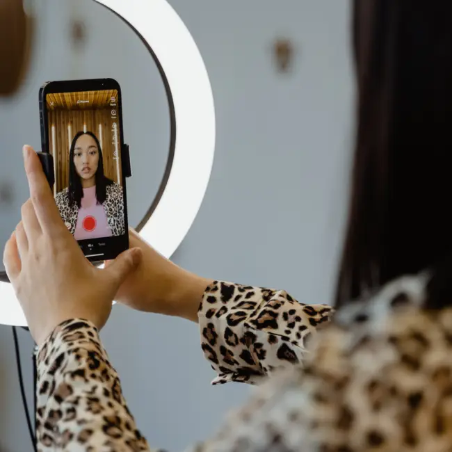 A person with long dark hair, wearing a leopard print shirt, holds a smartphone in front of a ring light, recording a video. The phone screen displays the person’s face, centered in the shot, with a red recording button visible on the screen.