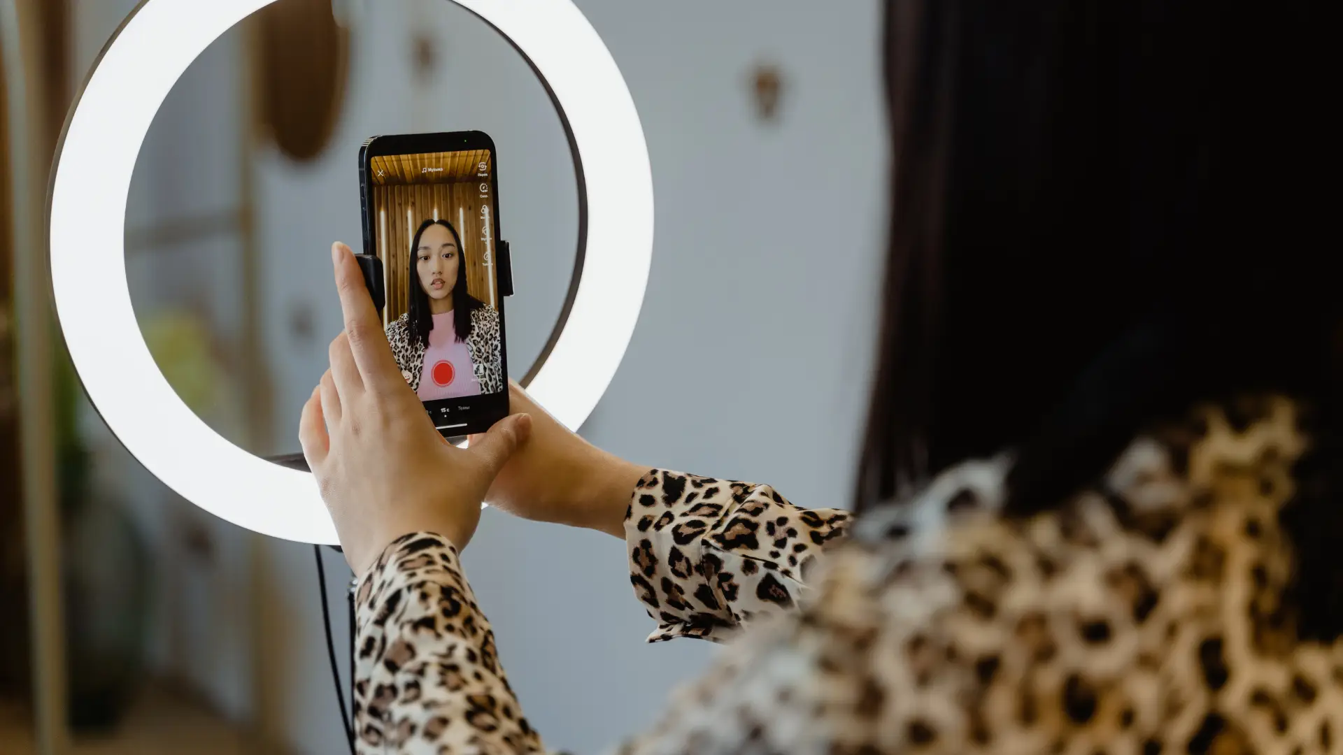 A person with long dark hair, wearing a leopard print shirt, holds a smartphone in front of a ring light, recording a video. The phone screen displays the person’s face, centered in the shot, with a red recording button visible on the screen.