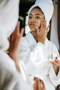 woman in a white robe and towel wrapped around her head, applying face cream while looking into a mirror, holding a small jar of skincare product.