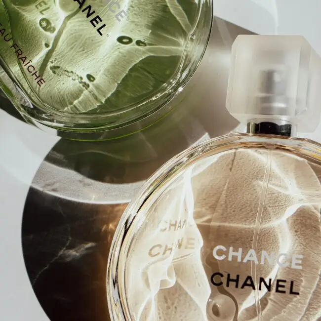 Two Chanel Chance perfume bottles, one in green (Eau Fraîche) and one in clear (Eau Vive), placed on a reflective surface, creating light and shadow patterns.