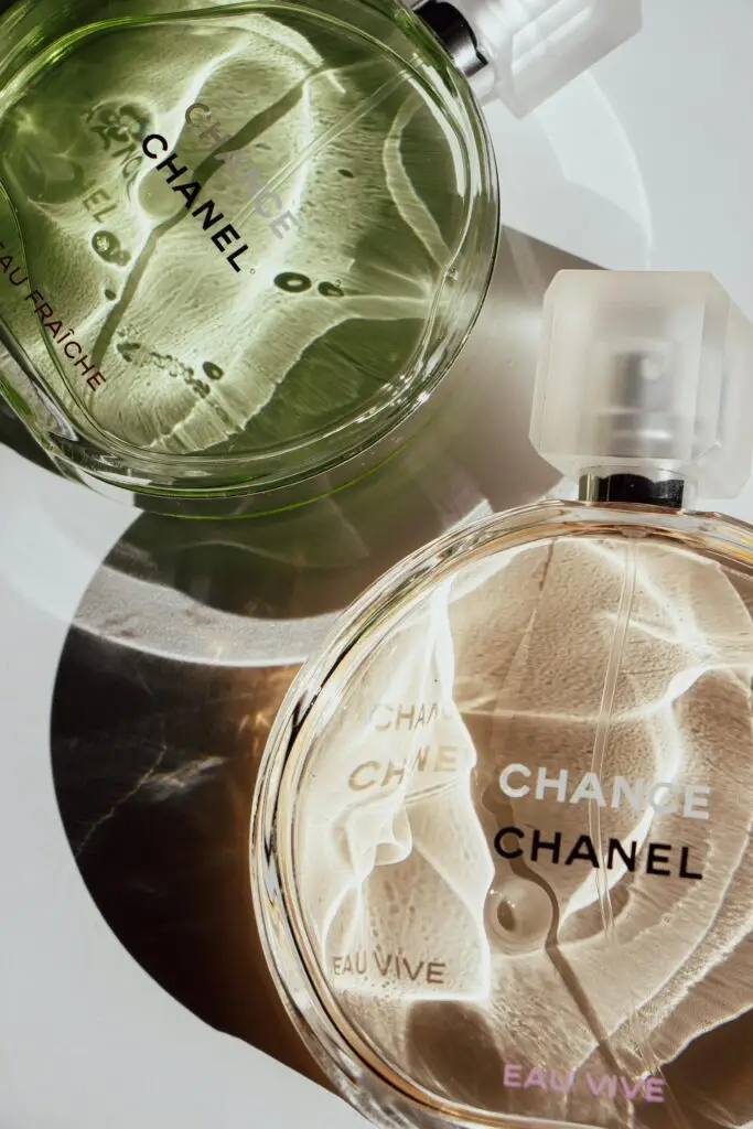 Two Chanel Chance perfume bottles, one in green (Eau Fraîche) and one in clear (Eau Vive), placed on a reflective surface, creating light and shadow patterns.